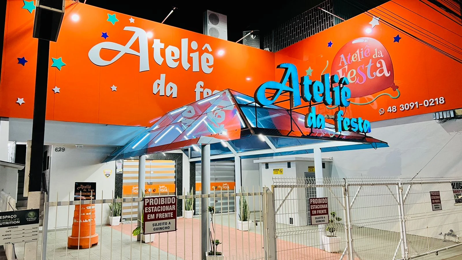 Fachada laranja do Ateliê da Festa fotografada à noite, com o nome da loja em destaque.